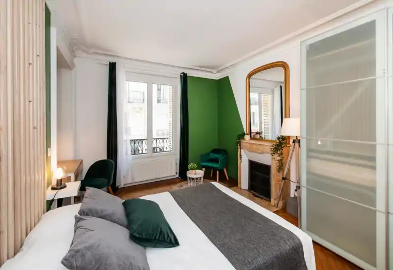 Bright, stylish bedroom with a large bed in the foreground, green accent wall, decorative fireplace and a mirror. The room is well lit with natural light and cozy textiles, ideal for rental photos.