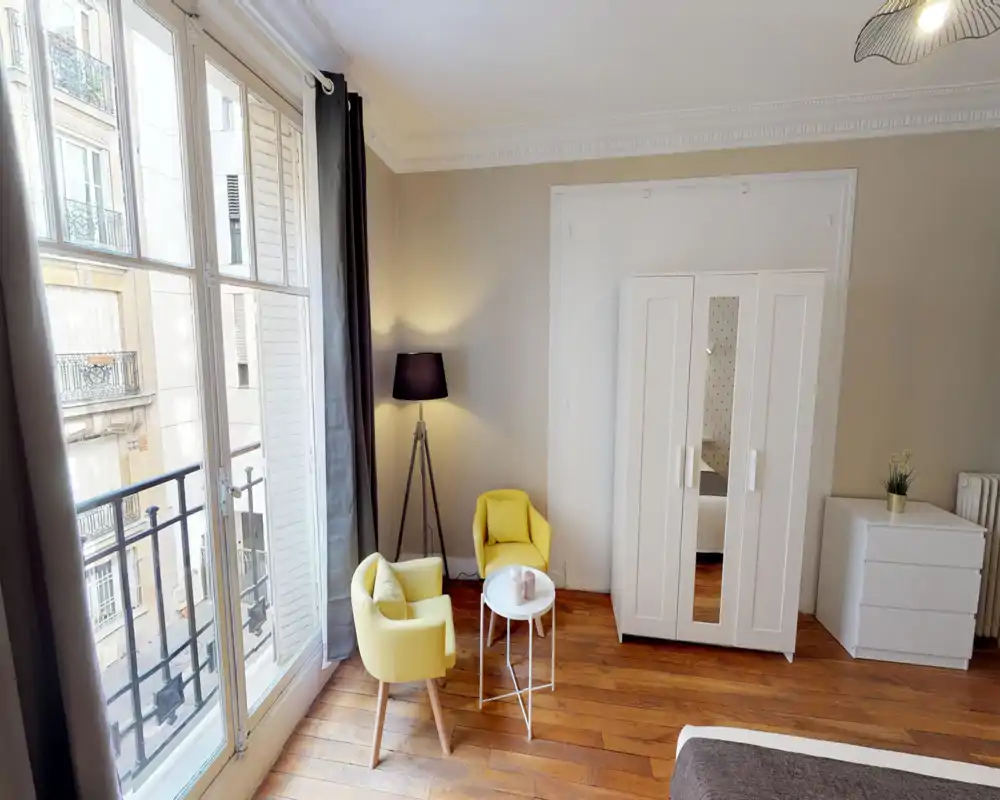 Bright, stylish bedroom corner with large French windows, two yellow armchairs and a wardrobe; partial view of the bed in the foreground and warm wooden floors.