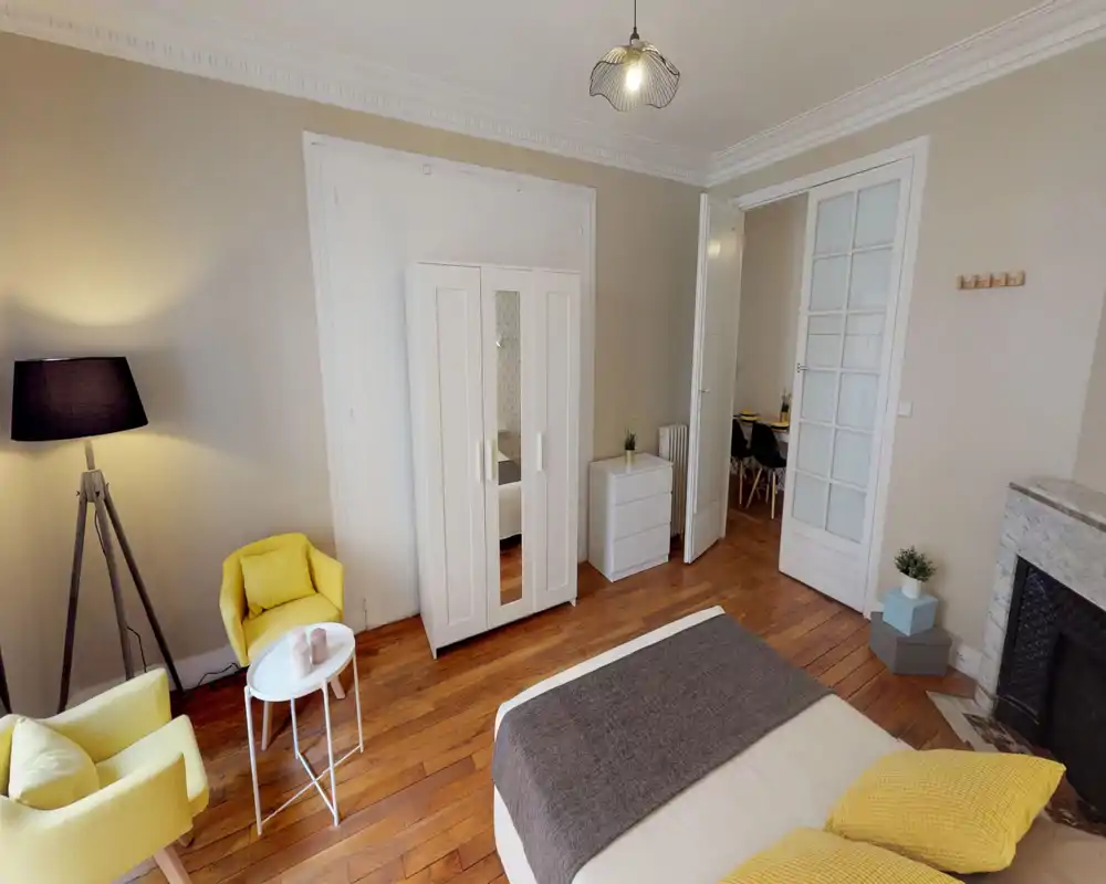 Bright, stylish bedroom with a large bed in the foreground, cozy yellow armchairs, and a white wardrobe. Warm wooden floors and tasteful lighting create a welcoming atmosphere ideal for rental listings.