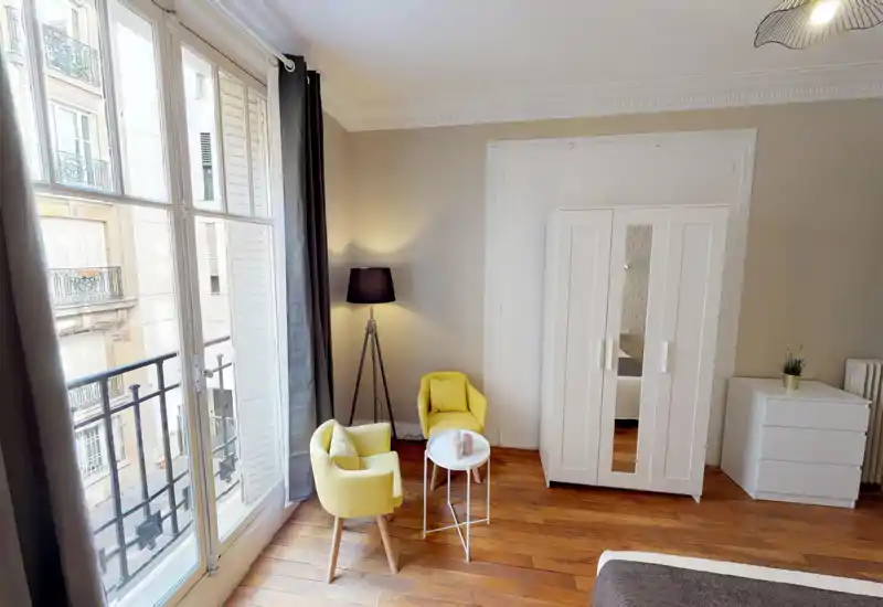 Bright, stylish bedroom corner with large French windows, two yellow armchairs and a wardrobe; partial view of the bed in the foreground and warm wooden floors.