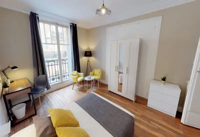 Bright, stylish bedroom with a double bed dressed in neutral linens and yellow accent cushions, a large window with natural light, a wardrobe and a small seating area — ideal for a cozy city stay.