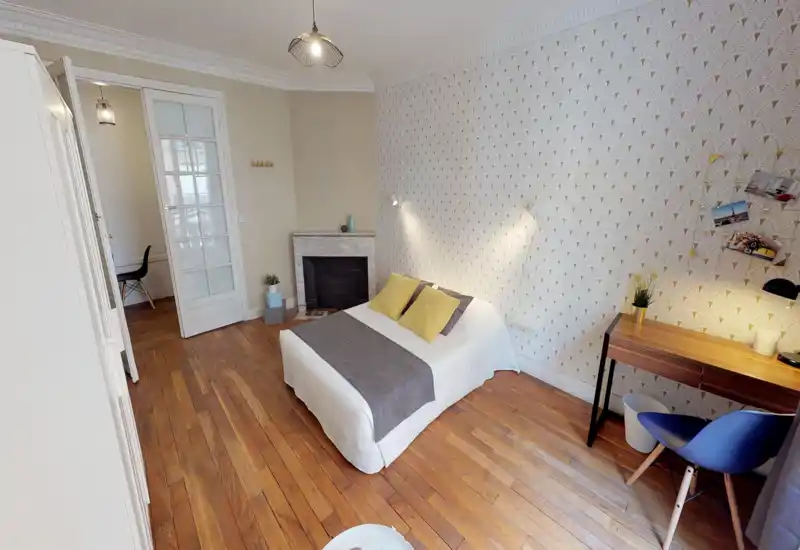 Bright, stylish bedroom with a centrally placed double bed, decorative pillows and a wooden floor. A small desk and chair near the window add workspace; patterned wallpaper and soft lighting give a cozy, modern feel.