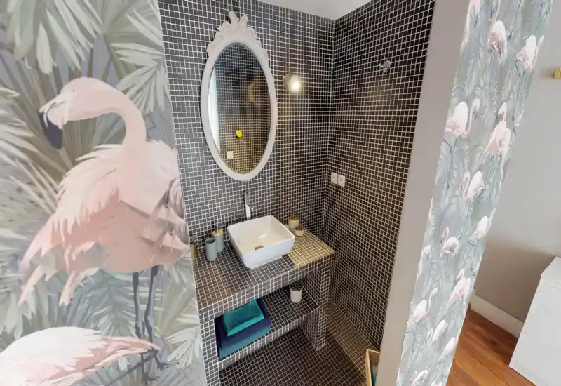 Angolo bagno compatto con piastrelle a mosaico scure, lavabo a bacinella e specchio ovale; carta da parati con fenicotteri per un tocco decorativo.