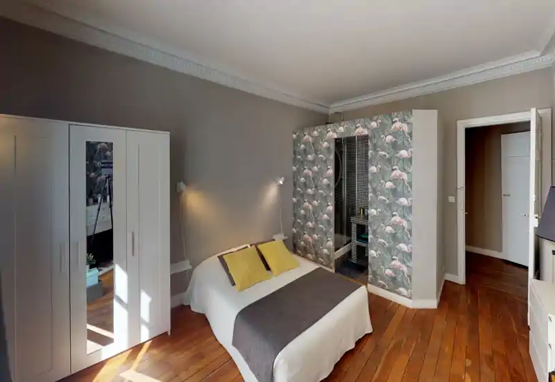 Camera da letto luminosa e di stile con letto matrimoniale, pavimento in legno caldo e carta da parati con fenicotteri attorno a un'area doccia integrata. Buona luce naturale, lampade vicino al letto e tessili accoglienti creano un'atmosfera invitante.