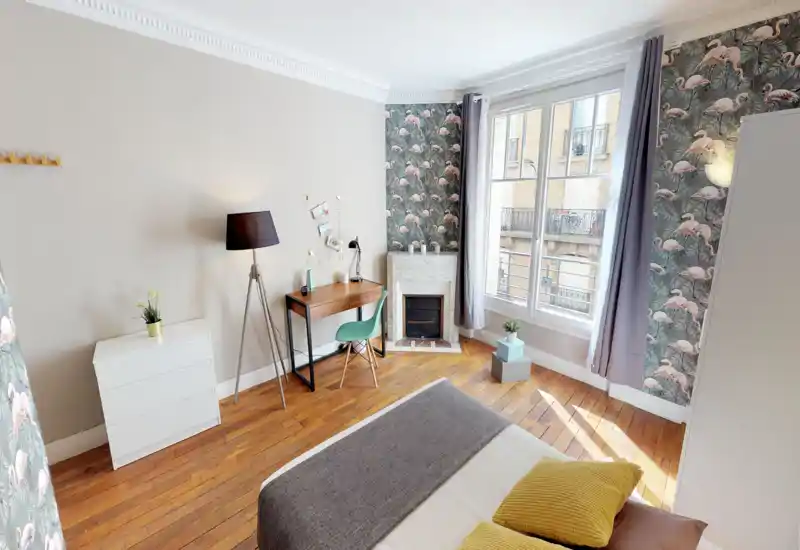 Camera da letto luminosa e ben arredata con pavimenti in legno, ampia finestra e letto rifatto; include una scrivania, carta da parati con fenicotteri e tessili accoglienti, perfetta per un soggiorno cittadino.