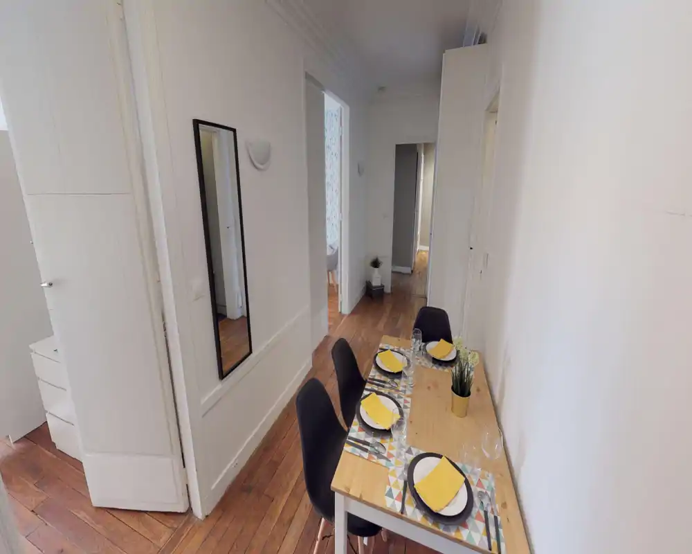 A narrow dining area with a wooden table set for four, modern black chairs and simple decorative touches like a small plant and yellow napkins. Bright, clean styling and hardwood floors create a warm, inviting space ideal for compact apartment listings.