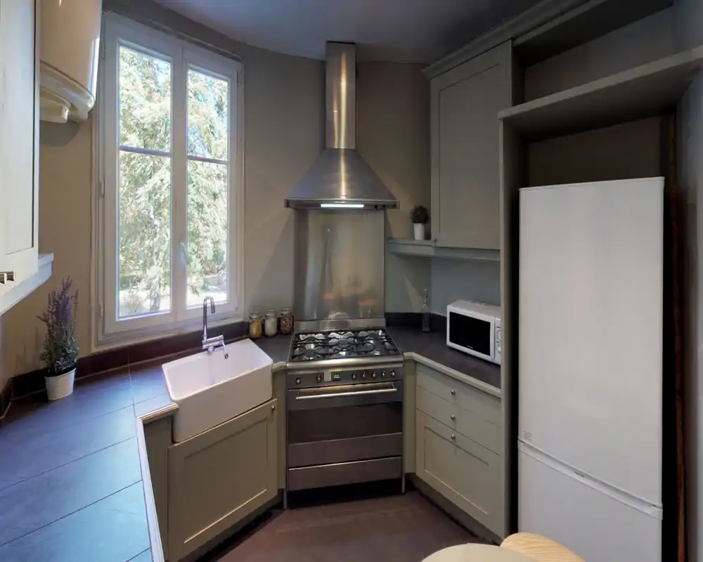 Bright, compact kitchen with a large window, farmhouse sink, gas range and stainless extractor hood. Neutral cabinetry, countertop workspace and a fridge-microwave setup create a practical, cozy cooking area.