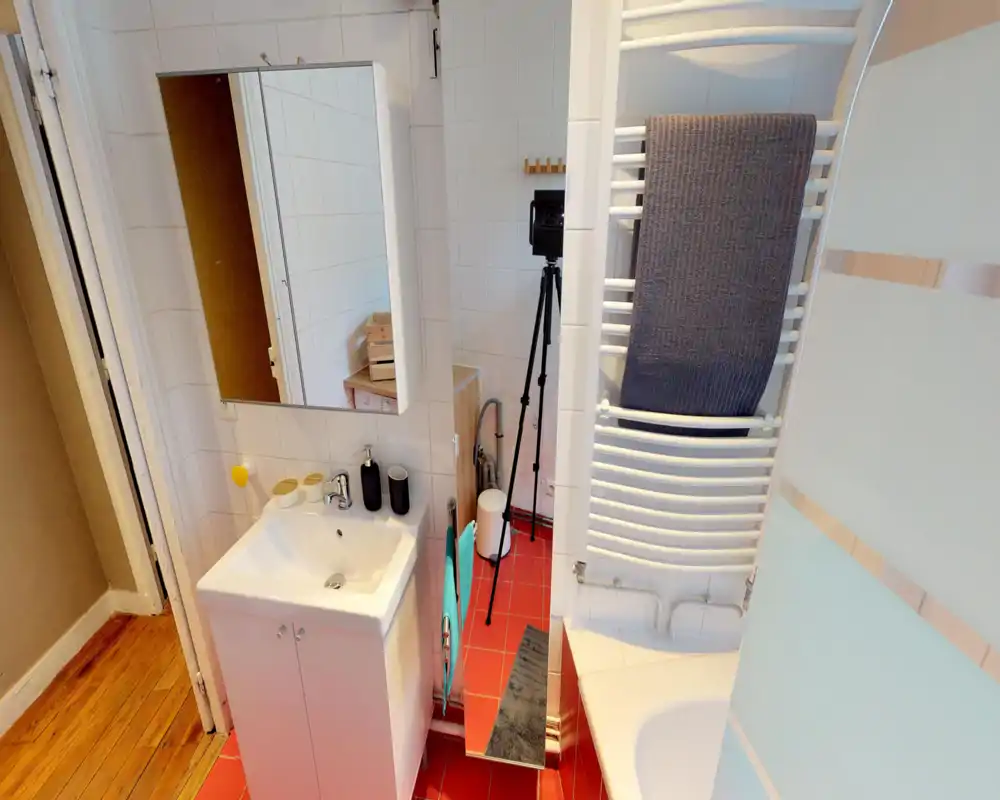 Clean, compact bathroom with a white sink and vanity, mirrored medicine cabinet, towel radiator with towels, and a bathtub with frosted glass screen. Bright red floor tiles add a pop of color.