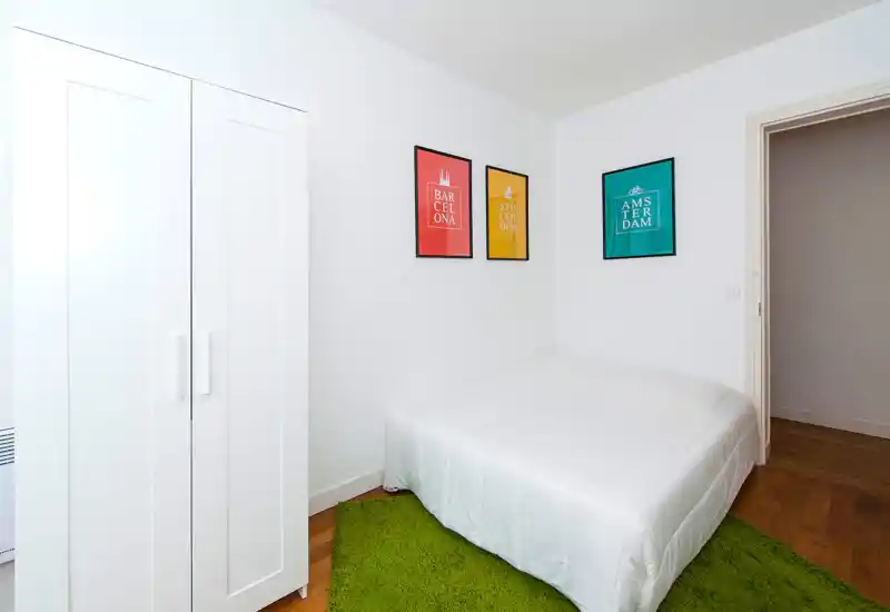 Bright, minimal bedroom with a white wardrobe, double bed with white bedding and colorful framed prints; wooden floor with a green rug adds warmth.
