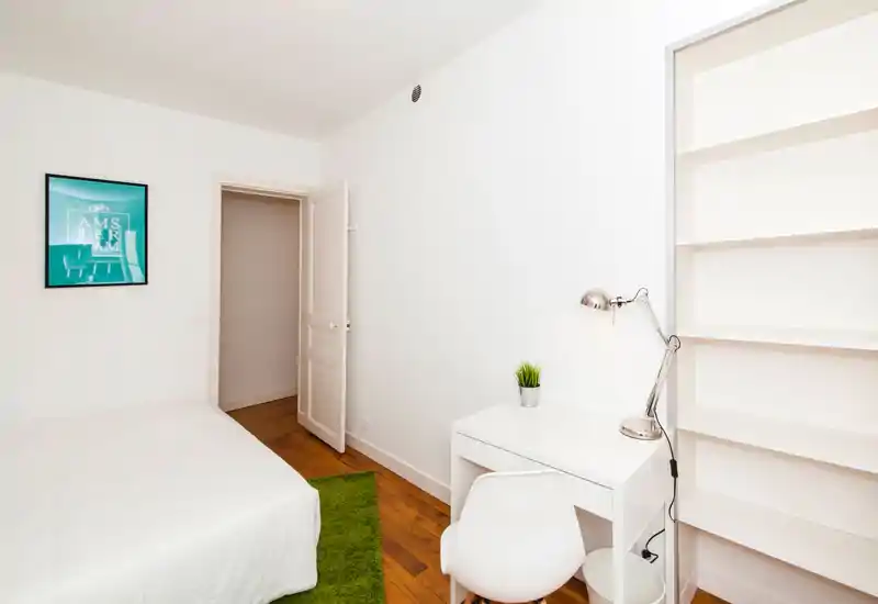 Bright, minimalist bedroom with a white bed, wooden floor, small green rug and a compact desk with shelving — ideal for a single occupant or student.