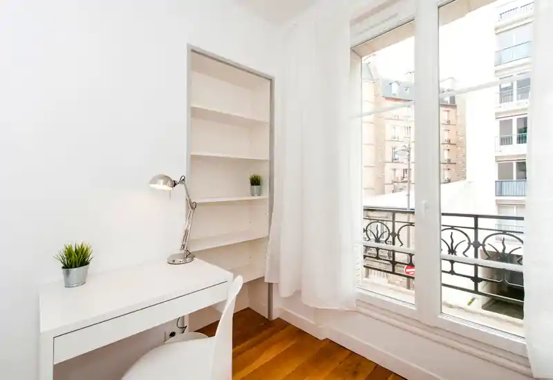 Bright, minimal corner with a white desk, lamp and shelving beside a large window overlooking the street — ideal as a small home office or study nook.