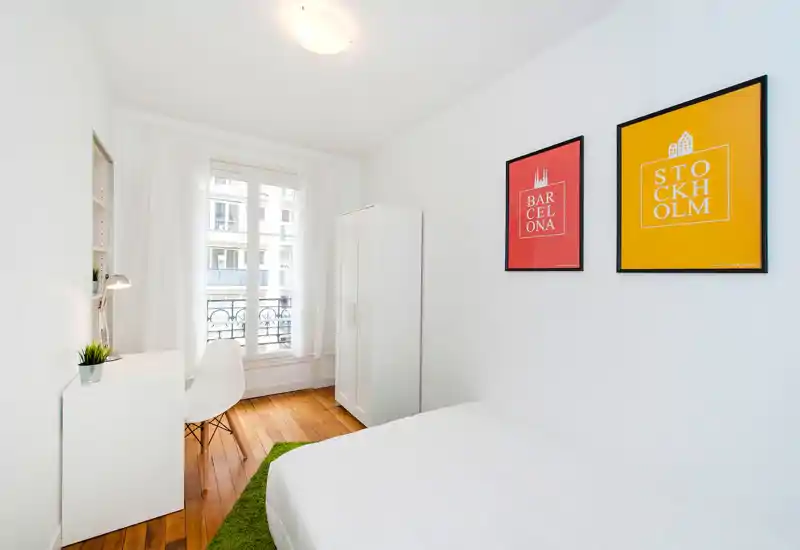 Bright, minimal bedroom with a white bed, wardrobe and desk by a large window. Clean Scandinavian-style décor with warm wooden floors and colorful framed prints.