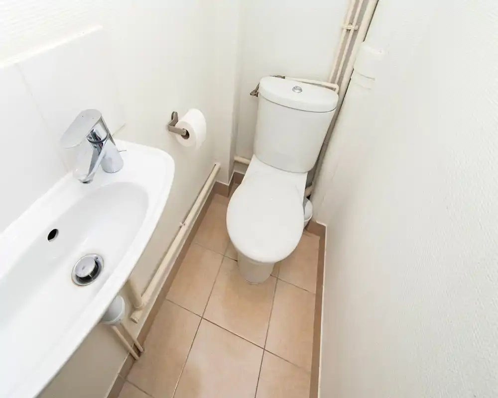 Compact, clean bathroom with a white toilet and wall-mounted sink, beige tiled floor and visible plumbing runs — bright and functional.