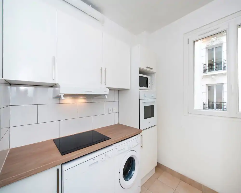Bright modern compact kitchen with white cabinets, wood countertop, induction hob and integrated washer/dishwasher; large window provides natural light.
