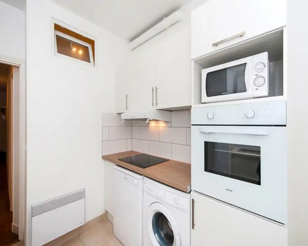 Compact, modern kitchen corner with white cabinets, built-in oven and microwave, induction hob and integrated appliances; clean wood countertop and tiled backsplash.