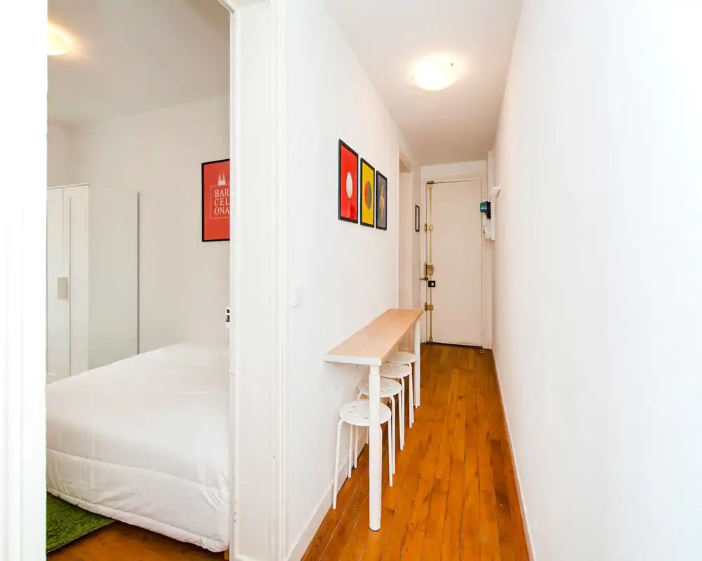 Bright, narrow entrance hallway with wooden floor, a slim console table and stools, wall art and the apartment door at the end.