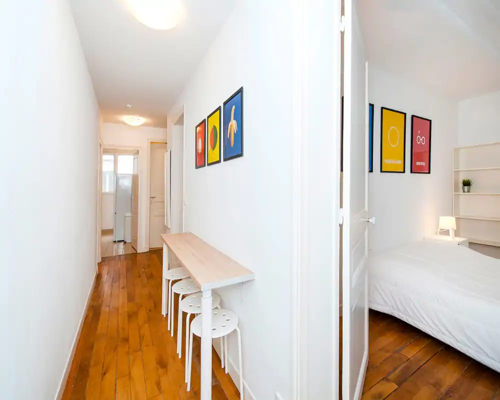 Bright, narrow entrance corridor with wooden floors, simple console table and stools; leads to a bedroom and kitchen area.