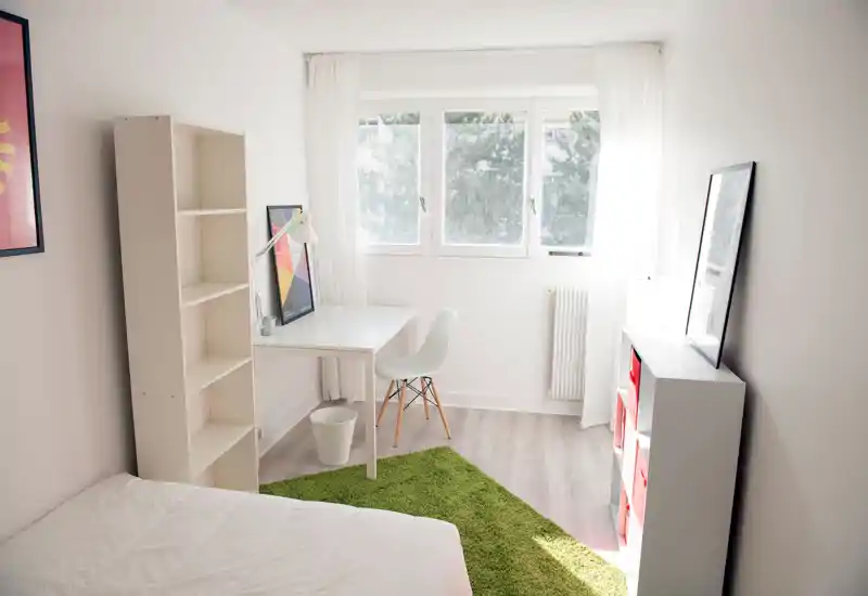 Chambre lumineuse et minimaliste avec lit blanc, petit bureau près d'une grande fenêtre et tapis vert; lignes épurées et lumière naturelle abondante, idéale pour étudiant ou personne seule.