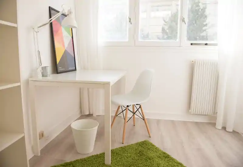 Coin bureau minimaliste et lumineux avec bureau blanc, chaise moderne, lampe et grande fenêtre laissant entrer la lumière naturelle — espace de travail aéré.