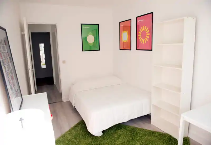 Chambre compacte et lumineuse avec un lit bien fait, linge blanc, affiches colorées et une étagère blanche vide — idéale pour une location moderne et minimaliste.