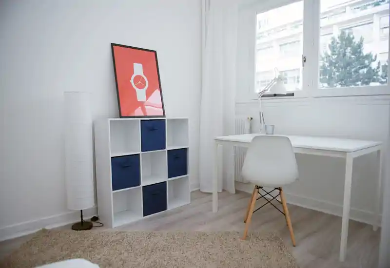 Rincón luminoso y minimalista de salón/estudio con escritorio blanco, silla, estantería en cubos y alfombra suave — ideal para una oficina en casa compacta o rincón de lectura.