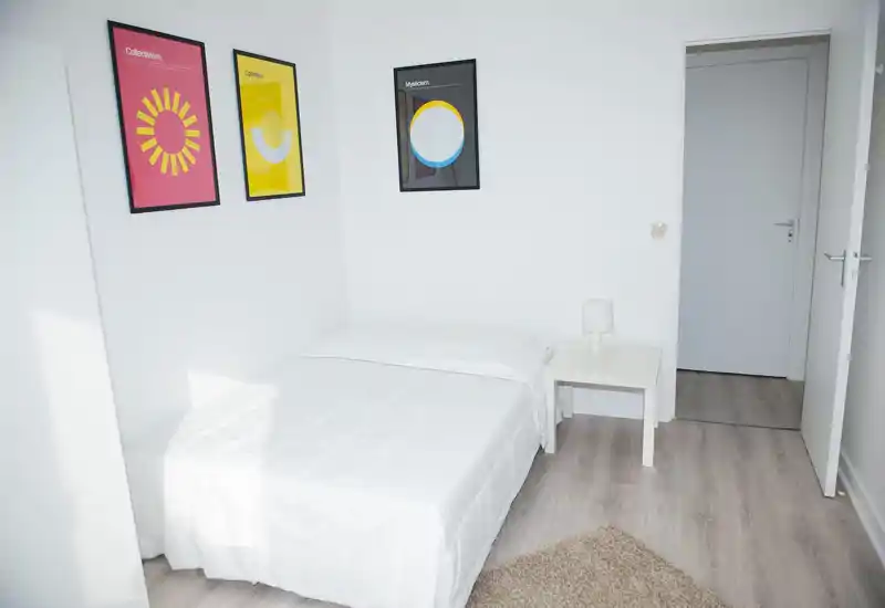 Dormitorio minimalista y luminoso con cama doble bien hecha, ropa de cama blanca, mesita con lámpara y tres cuadros coloridos en la pared. Diseño nórdico limpio, buena luz natural y suelos de madera claros — ideal para un anuncio.