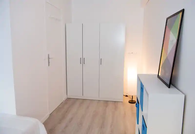 Coin de chambre minimaliste avec une armoire blanche à trois portes, une lampe sur pied et une étagère avec bacs de rangement bleus ; tons neutres et sol propre pour une ambiance calme.