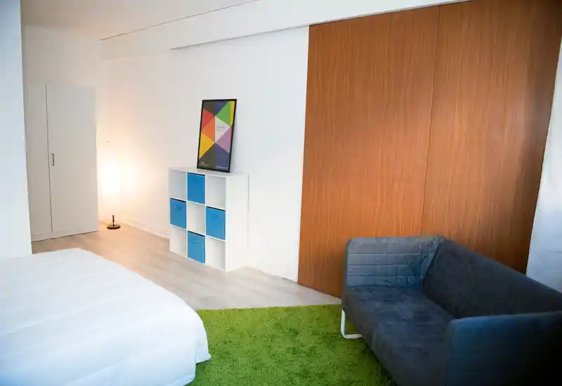 Chambre moderne et lumineuse avec lit blanc visible, tapis vert et étagères cubiques ; penderie coulissante en bois chaleureux et petit canapé gris pour une ambiance minimaliste et fraîche.
