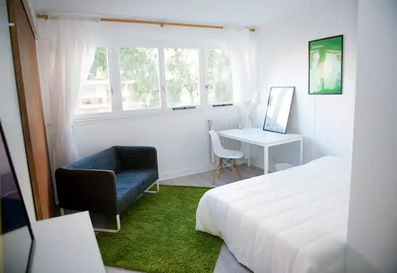 Chambre lumineuse et minimaliste avec un grand lit, un petit canapé et un bureau près de la fenêtre. La lumière naturelle inonde la pièce grâce aux grandes fenêtres, offrant une atmosphère fraîche et aérée idéale pour la location.