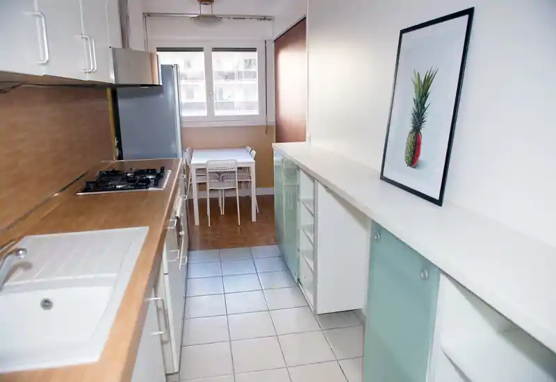 Cuisine en couloir moderne et lumineuse avec plans de travail en bois, plaque à gaz encastrée, évier et petit coin repas près de la fenêtre. Style épuré et minimaliste avec placards blancs et tableau décoratif.