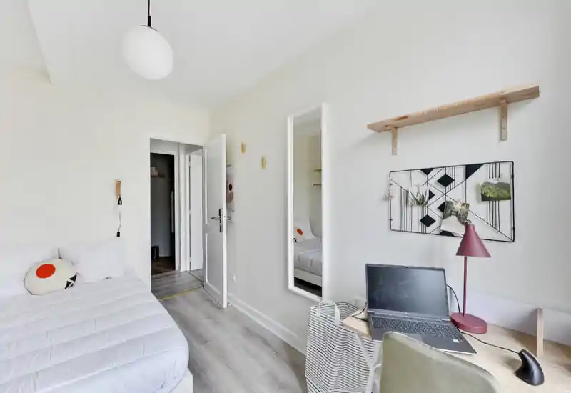 Chambre simple moderne et lumineuse avec un lit bien fait, un miroir mural et un petit espace de travail avec un ordinateur portable—idéale pour un étudiant ou un professionnel. Décor épuré et lumière naturelle agréable.