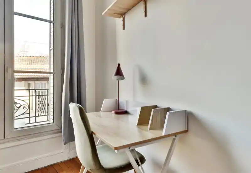 Coin bureau lumineux et minimaliste avec un bureau en bois, une chaise rembourrée et une lampe de bureau rouge près d'une grande fenêtre qui inonde la pièce de lumière naturelle.
