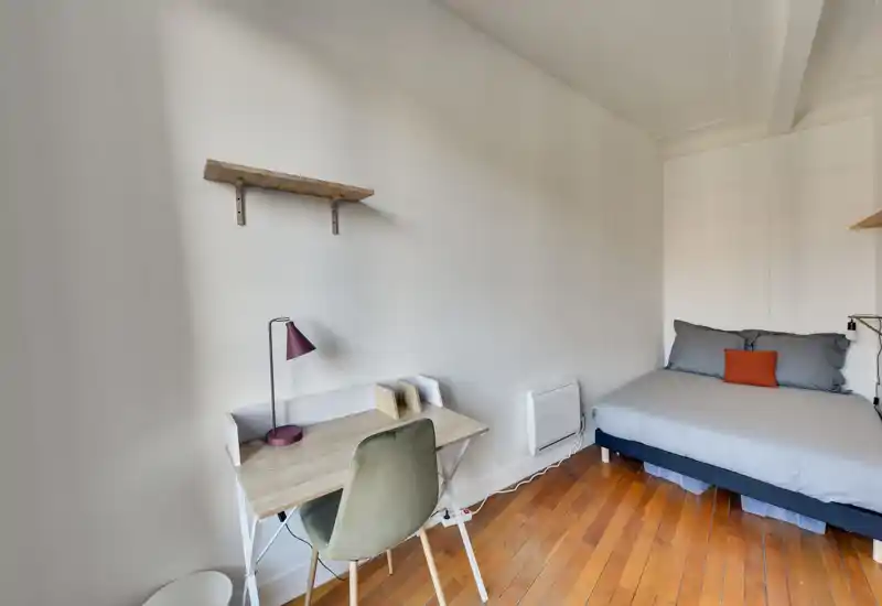 Chambre compacte et lumineuse avec un lit double en linge gris et un coussin orange contrastant, ainsi qu'un petit bureau en bois et une chaise — idéale pour travailler et se reposer.