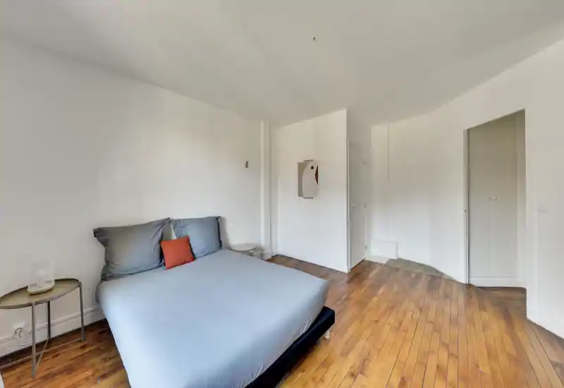 Chambre lumineuse et minimaliste avec un lit double bien fait en linge gris, un sol en bois chaleureux et une petite table de chevet. L'espace paraît aéré et épuré, idéal pour des photos de location mettant en valeur calme et volume.