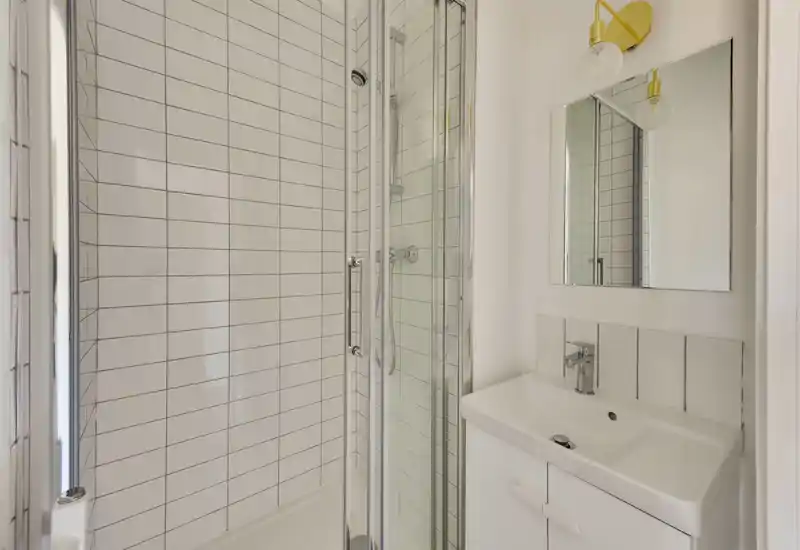 Salle de bain compacte et moderne avec douche carrelée, porte vitrée et meuble vasque blanc avec miroir — lumineux et propre.