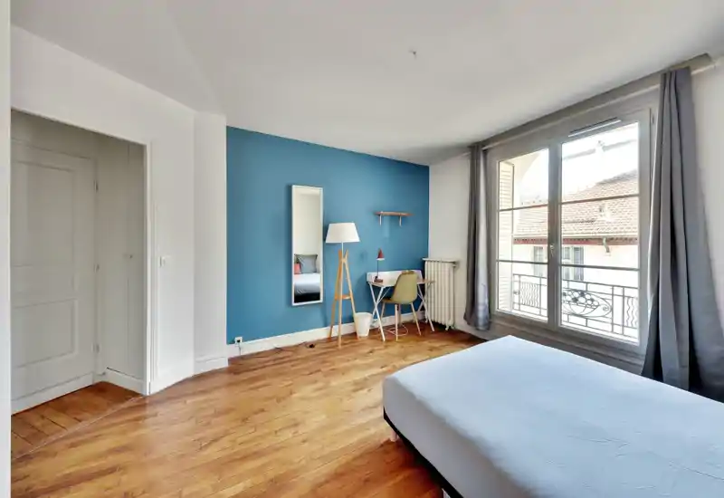 Chambre lumineuse et moderne avec une grande fenêtre, un mur bleu d'accent et un sol en bois ; matelas visible au premier plan. Un petit bureau, une lampe sur pied et un miroir apportent une touche fonctionnelle pour les locations.