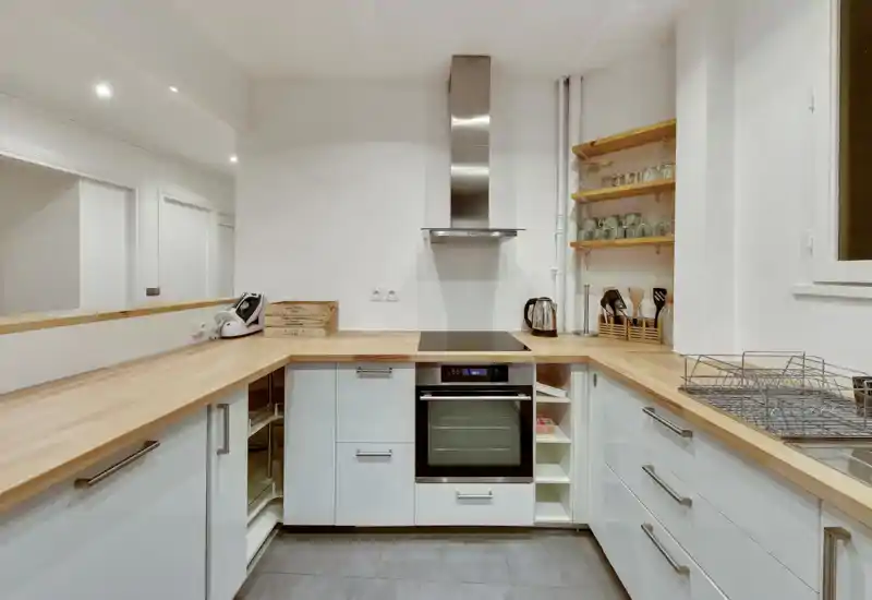 Cuisine moderne et compacte avec placards blancs, plans de travail en bois, four encastré et plaque à induction, étagères ouvertes avec verrerie et rangement d'ustensiles soigné — espace lumineux et fonctionnel adapté aux locations courte durée.