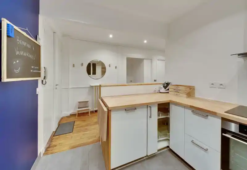 Cuisine moderne ouverte avec plans de travail en bois, placards blancs et plaque à induction. Espace lumineux et bien agencé avec un sol en bois visible à côté.