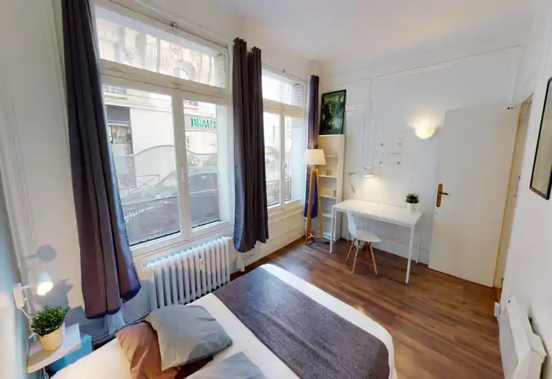 Bright, modern bedroom with a large bed in the foreground, tall windows with dark curtains, a desk and shelving by the wall, and warm wooden floors — ideal for a city apartment.