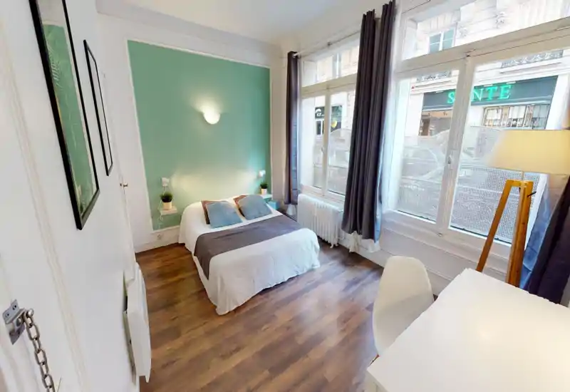 Bright, well-staged bedroom with a double bed centered against a mint-green accent wall, bedside shelves with plants, large windows letting in natural light, and a small desk — ideal for rental listing photos.