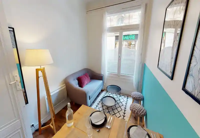 Compact, stylish living area with a small sofa, decorative cushions, patterned rug and two nesting coffee tables by a large window offering natural light.
