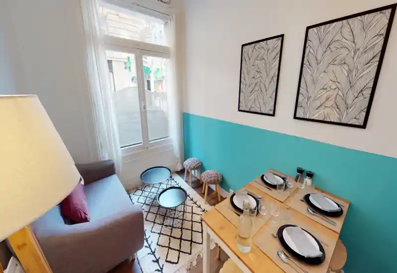 Bright, well-styled living area with a compact sofa, patterned rug and two nesting coffee tables next to a window; a small dining setup is visible on the right, making the space ideal for a cozy city stay.
