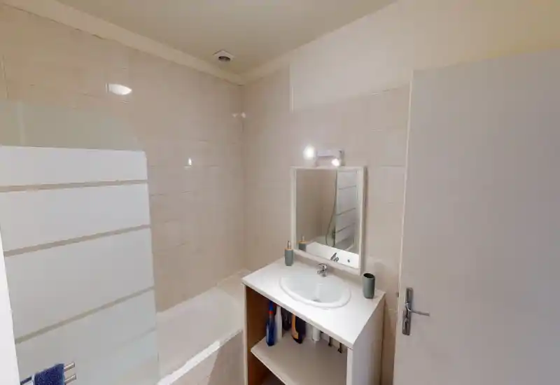 Compact tiled bathroom with bathtub and a vanity sink under a mirror. Neutral beige tiles, simple lighting and modest storage make it practical for short stays.