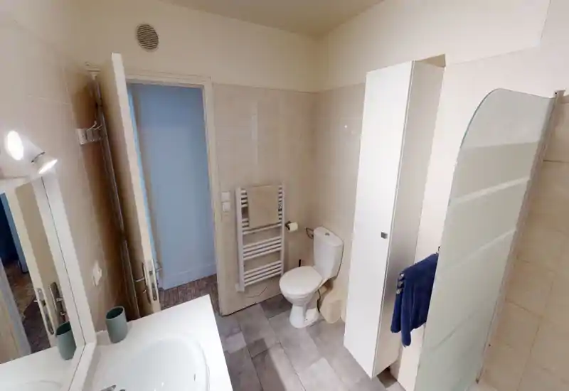 Clean, compact bathroom with a sink and mirror on the left, a toilet and towel radiator in the center, and a shower with frosted screen on the right. Neutral tiles and modern fixtures create a tidy, functional space.
