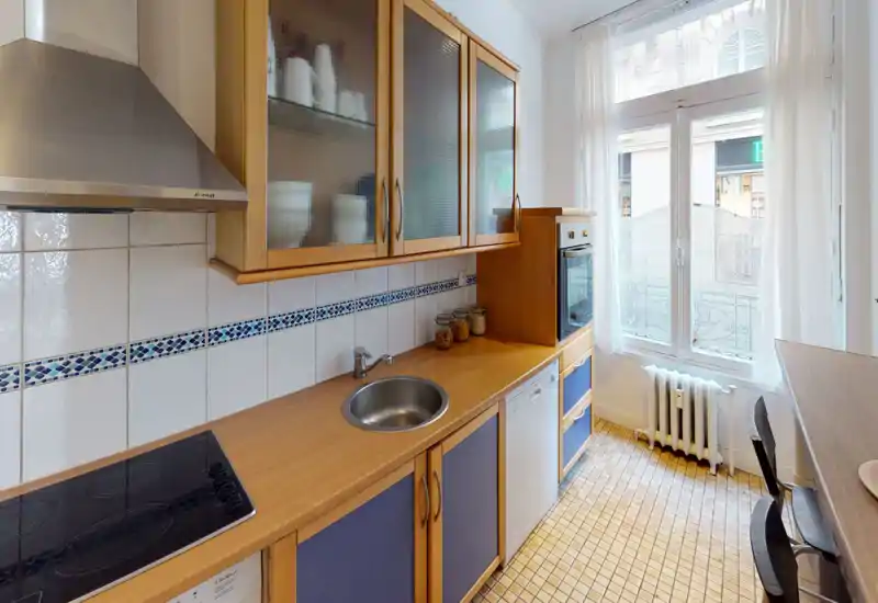 Bright, compact kitchen with wooden cabinets, built-in oven, ceramic hob and a round sink. Large window provides natural light and a cozy tiled floor adds character — ideal for city apartment listings.