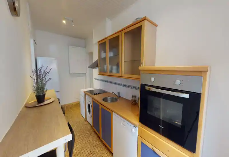 Compact fitted kitchen with wooden cabinets, built-in oven, hob, dishwasher and a small prep counter; warm tones and practical layout ideal for a city apartment.