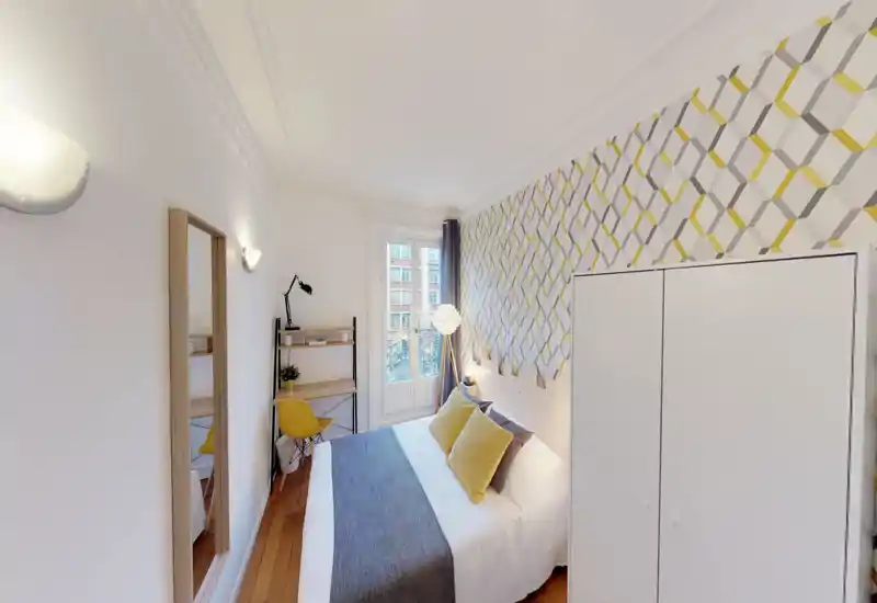 Camera da letto luminosa e ben arredata con letto matrimoniale, cuscini decorativi e parete a motivo. La luce naturale dalla finestra francese e l’illuminazione calda rendono l’ambiente accogliente; scrivania e armadio completano la stanza.