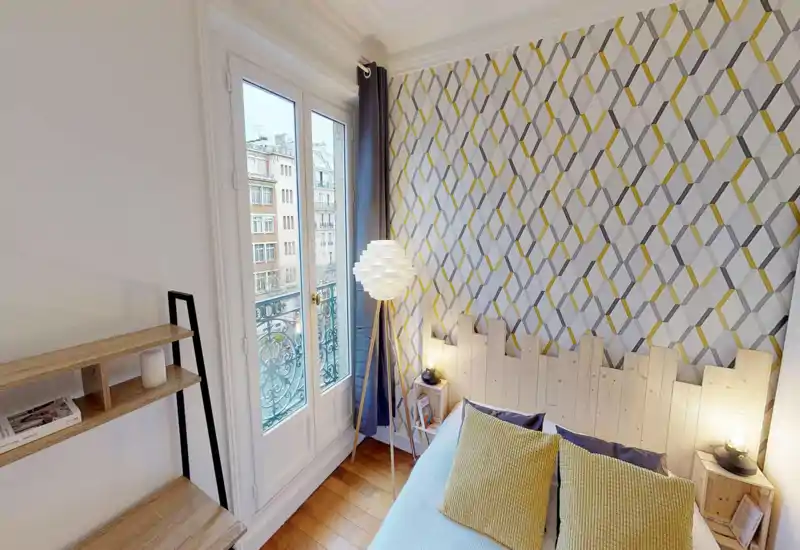 Camera da letto luminosa e moderna con carta da parati geometrica, testiera in legno e cuscini senape; luce naturale dalla porta-finestra e lampade soffuse rendono l'ambiente accogliente.
