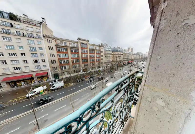 Piccolo balcone che si affaccia su un viale cittadino trafficato, con ringhiera in ferro battuto decorativa e vista sulla strada sottostante.