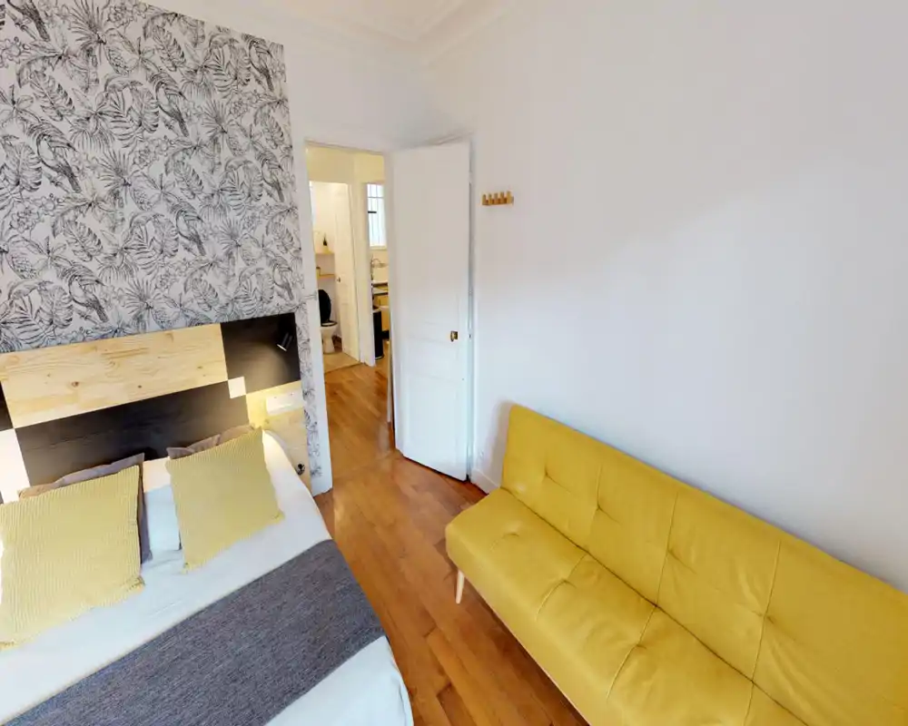Dormitorio luminoso y con estilo con pared decorativa, cabecero de madera y un sofá amarillo. La cama está bien tendida con cojines a juego y el suelo de madera aporta calidez.